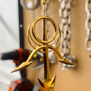 May include: A gold-colored pendant necklace featuring a stylized design with a circle and three arrow-like points. The pendant is hanging from a black leather cord.