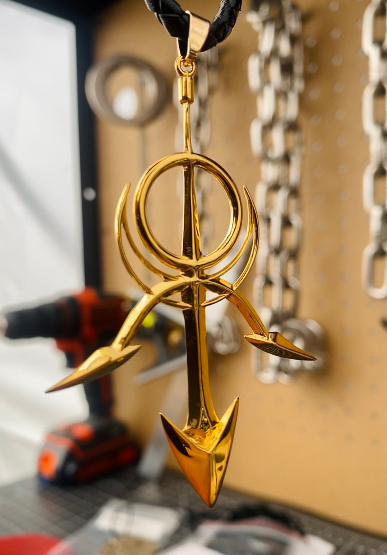 May include: A gold-colored pendant necklace featuring a stylized design with a circle and three arrow-like points. The pendant is hanging from a black leather cord.
