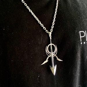 May include: A silver chain necklace with a pendant featuring a stylized arrow design. The arrow is pointed upwards and has a circular element at the top.