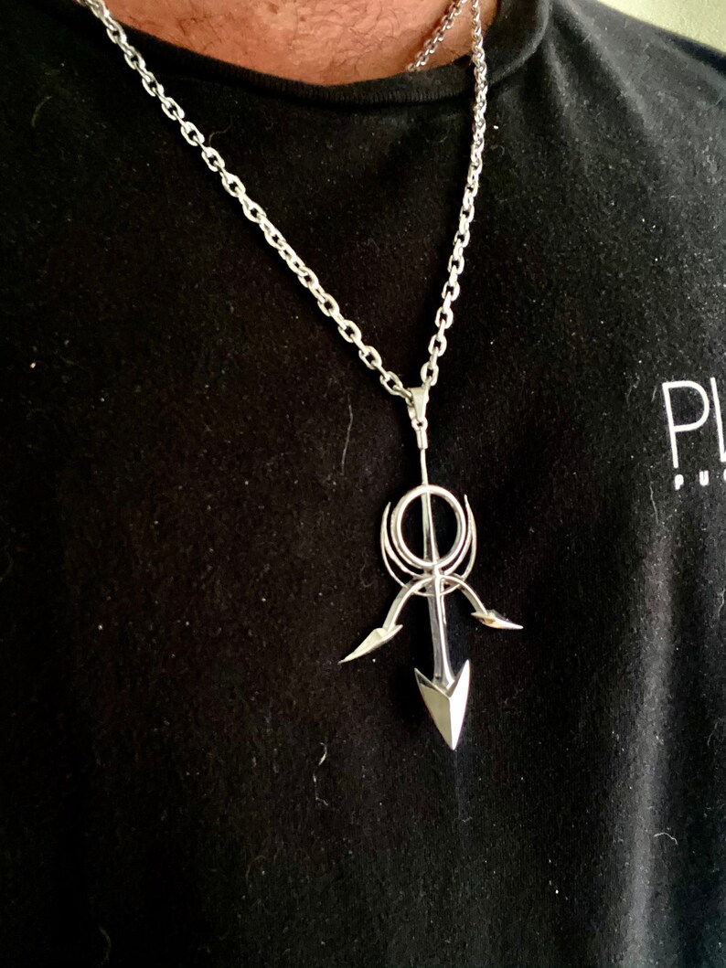 May include: A silver chain necklace with a pendant featuring a stylized arrow design. The arrow is pointed upwards and has a circular element at the top.
