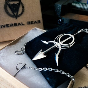 May include: A silver necklace with a large, intricate pendant featuring a stylized arrow design. The necklace is displayed in a black velvet pouch and a brown box with the text "UNIVERSAL BEAR" printed on the top.