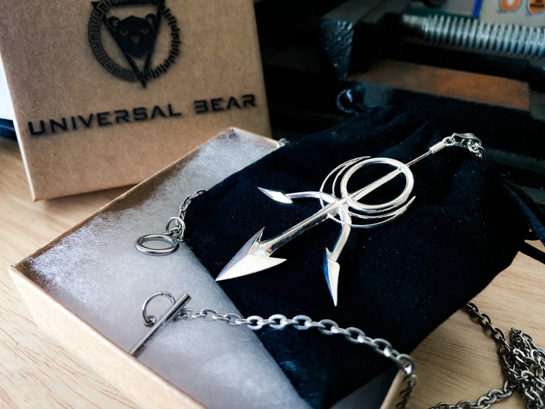 May include: A silver necklace with a large, intricate pendant featuring a stylized arrow design. The necklace is displayed in a black velvet pouch and a brown box with the text "UNIVERSAL BEAR" printed on the top.