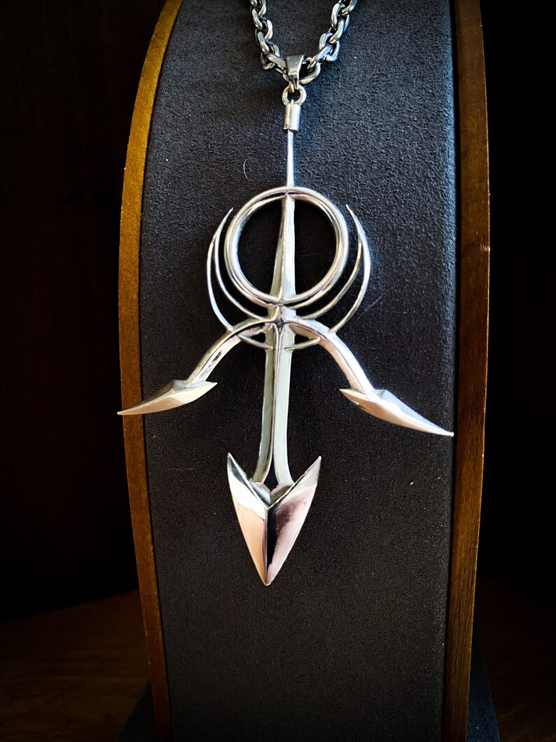 May include: A silver pendant necklace with a geometric design featuring a circle, a cross, and two curved lines. The pendant is attached to a silver chain.