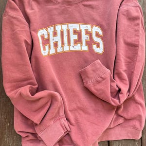 May include: A red sweatshirt with the word "CHIEFS" printed in white with a yellow outline. The sweatshirt is made of a soft, comfortable fabric.