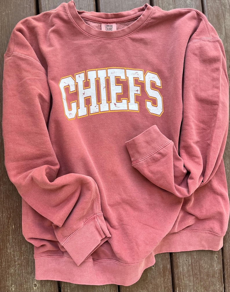 May include: A red sweatshirt with the word "CHIEFS" printed in white with a yellow outline. The sweatshirt is made of a soft, comfortable fabric.