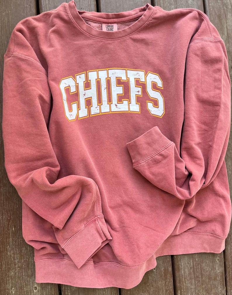 May include: A red sweatshirt with the word "CHIEFS" printed in white with a yellow outline. The sweatshirt is folded and laid on a wooden surface.