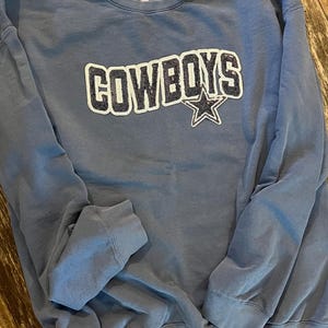May include: A dusty blue sweatshirt with the word "COWBOYS" in a white outlined, sequined font. A sequined star is below the word. The sweatshirt is made of a soft material and has a crew neck.