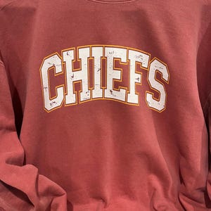 May include: A maroon long-sleeve t-shirt with a white distressed graphic of the word "CHIEFS" in an arched font with an orange outline.