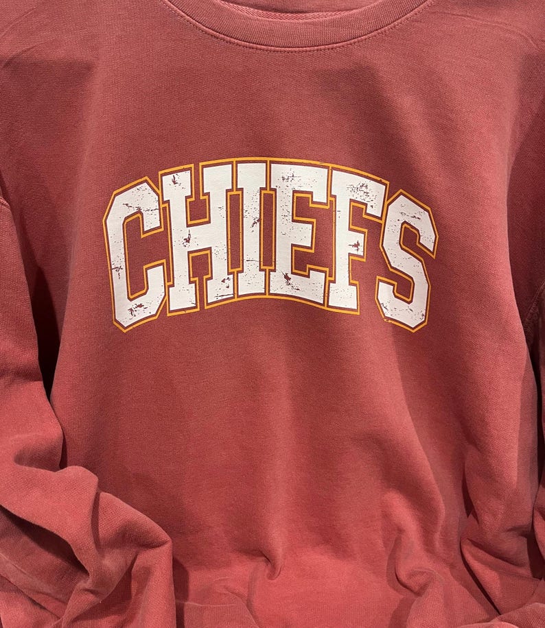 May include: A maroon long-sleeve t-shirt with a white distressed graphic of the word "CHIEFS" in an arched font with an orange outline.