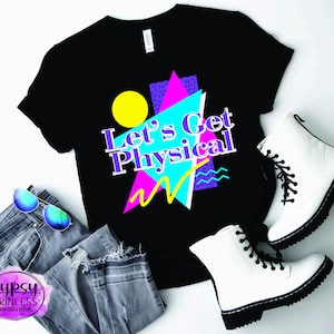 Può includere: Maglietta nera con un design grafico che presenta un cerchio giallo, forme geometriche rosa, blu e viola, e il testo "Let's Get Physical".
