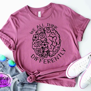 以下が含まれることがあります： 花で囲まれた脳の黒いグラフィックが描かれたピンクのTシャツ。Tシャツには「We all think differently」という文字が印刷されています。
