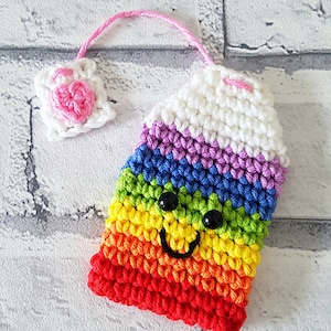 May include: A rainbow crocheted tea cozy with a smiling face and black eyes. The cozy is attached to a pink crocheted heart with a pink string.