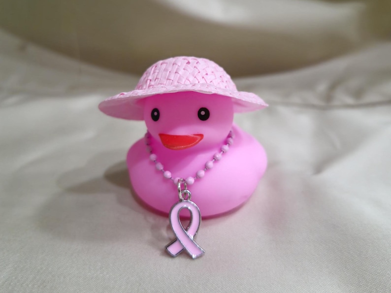 May include: A pink rubber duck wearing a pink straw hat and a pink ribbon necklace. The ribbon is a symbol of breast cancer awareness.