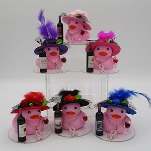 May include: Pink rubber duck figurines wearing hats with feathers and holding wine bottles and glasses. Each duck has a pink ribbon and a pearl necklace. The ducks are displayed on clear circular stands.