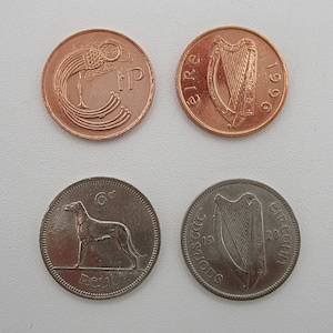 May include: Four Irish coins are displayed, two copper and two silver. The copper coins feature a harp and a bird design. The silver coins show a greyhound and a harp. The coins are from the Republic of Ireland.
