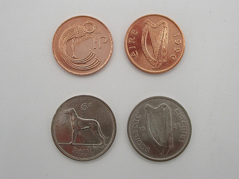 May include: Four Irish coins are displayed, two copper and two silver. The copper coins feature a harp and a bird design. The silver coins show a greyhound and a harp. The coins are from the Republic of Ireland.
