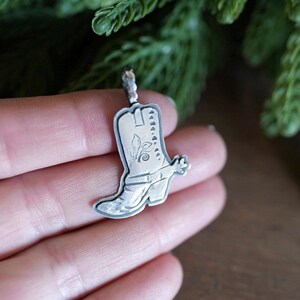 May include: A silver-toned metal pendant shaped like a cowboy boot. The pendant features engraved detailing on the surface, including a small floral motif on the shaft and a spur attached to the heel. It has a small loop at the top for hanging.