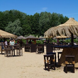 K&ouml;nnte beinhalten: Eine Strandbar-Szene mit mehreren strohgedeckten Strukturen. Tische und Hocker stehen auf dem Sand, w&auml;hrend Menschen den sonnigen Tag genie&szlig;en. Die Bar hat eine rustikale, tropische Atmosph&auml;re.