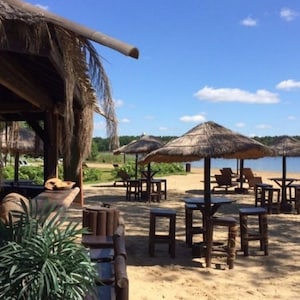 K&ouml;nnte beinhalten: Eine sonnige Strandszene mit Strohschirmen, Tischen und Hockern. Links befindet sich eine Holzkonstruktion mit Strohdach. Der Strand hat Liegest&uuml;hle und einen See im Hintergrund. Der Himmel ist blau mit vereinzelten Wolken.