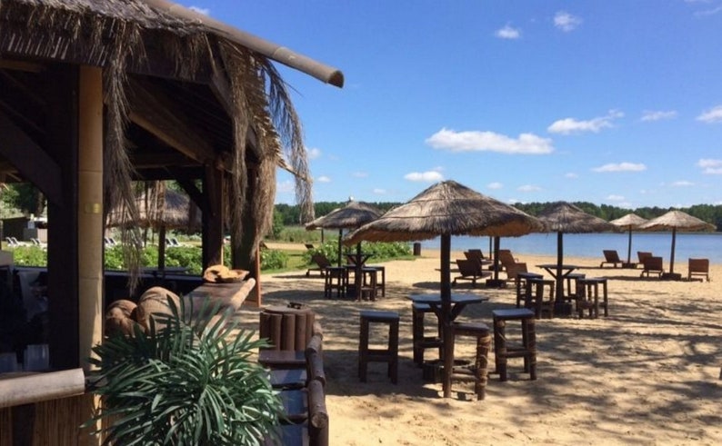 K&ouml;nnte beinhalten: Eine sonnige Strandszene mit Strohschirmen, Tischen und Hockern. Links befindet sich eine Holzkonstruktion mit Strohdach. Der Strand hat Liegest&uuml;hle und einen See im Hintergrund. Der Himmel ist blau mit vereinzelten Wolken.