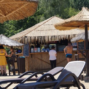 Puede incluir: Un bar con techo de paja, un mostrador de madera y taburetes. Hay gente sentada en el bar y otros en la cola. Tambi&eacute;n hay tumbonas en primer plano.