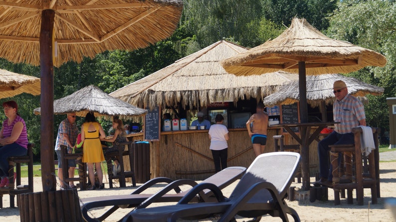 Puede incluir: Un bar con techo de paja, un mostrador de madera y taburetes. Hay gente sentada en el bar y otros en la cola. Tambi&eacute;n hay tumbonas en primer plano.
