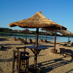 Puede incluir: Una sombrilla de paja ofrece sombra en una playa de arena con tumbonas de madera y una mesa y un taburete de madera. La playa est&aacute; junto a una masa de agua.