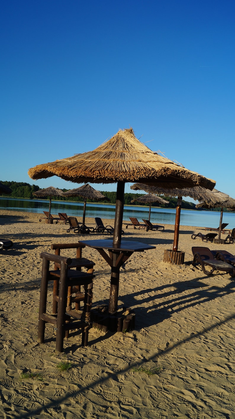 Puede incluir: Una sombrilla de paja ofrece sombra en una playa de arena con tumbonas de madera y una mesa y un taburete de madera. La playa est&aacute; junto a una masa de agua.