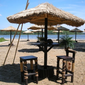 Puede incluir: Una escena de playa con dos taburetes de bar de madera y una mesa bajo una sombrilla de paja. La sombrilla est&aacute; hecha de paja tejida y tiene un marco de madera. La mesa est&aacute; hecha de madera y tiene un acabado marr&oacute;n. Los taburetes est&aacute;n hechos de madera y tienen un acabado natural. La playa es arenosa y hay un lago en el fondo.