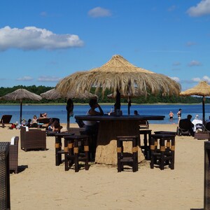 Puede incluir: Un bar de playa con sombrillas de paja y muebles de mimbre. La gente est&aacute; sentada en mesas y disfrutando del d&iacute;a soleado.
