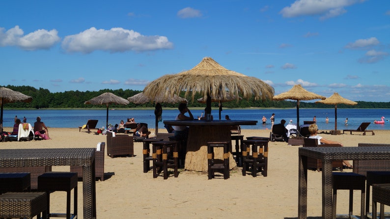 Puede incluir: Un bar de playa con sombrillas de paja y muebles de mimbre. La gente est&aacute; sentada en mesas y disfrutando del d&iacute;a soleado.