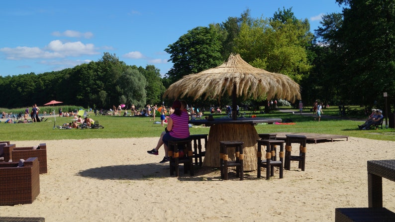 Puede incluir: Un bar tiki con techo de paja, una mesa de madera y sillas en una playa de arena. El bar est&aacute; rodeado de &aacute;rboles y hay gente en el fondo.
