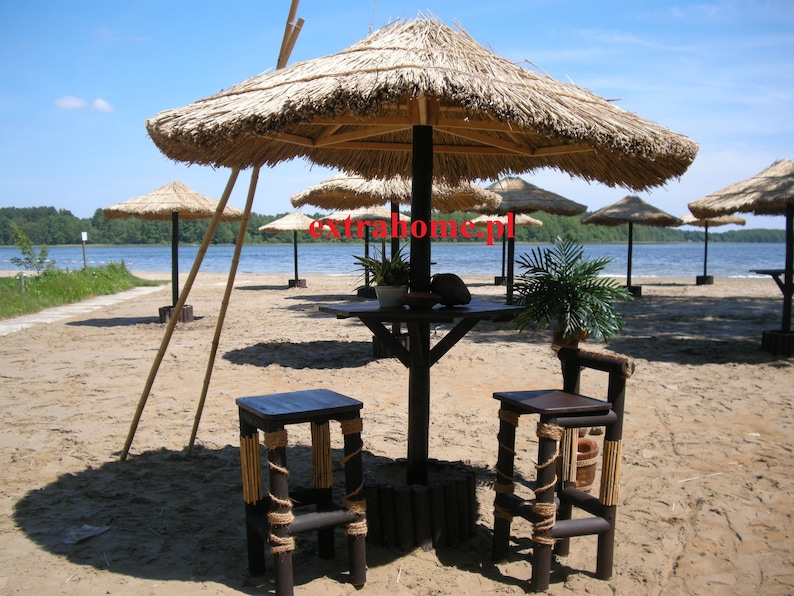 K&ouml;nnte beinhalten: Eine Strandszene mit mehreren Strohschirmen, die Schatten &uuml;ber Tischen und Hockern spenden. Die M&ouml;bel sind aus dunklem Holz und Seil gefertigt und stehen an einem Sandstrand mit einem See im Hintergrund. Der Text "extrahome.pl" ist sichtbar.