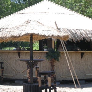 K&ouml;nnte beinhalten: Eine tropisch gestaltete Outdoor-Bar mit Strohdach und Bambusakzenten. Die Bar verf&uuml;gt &uuml;ber einen kleinen Sonnenschirm, einen Tisch und Hocker. Eine Leiter lehnt an der Bar. Die Bar ist von B&auml;umen und Gr&uuml;n umgeben.