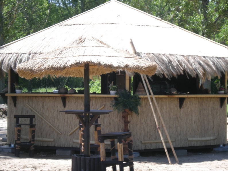 K&ouml;nnte beinhalten: Eine tropisch gestaltete Outdoor-Bar mit Strohdach und Bambusakzenten. Die Bar verf&uuml;gt &uuml;ber einen kleinen Sonnenschirm, einen Tisch und Hocker. Eine Leiter lehnt an der Bar. Die Bar ist von B&auml;umen und Gr&uuml;n umgeben.