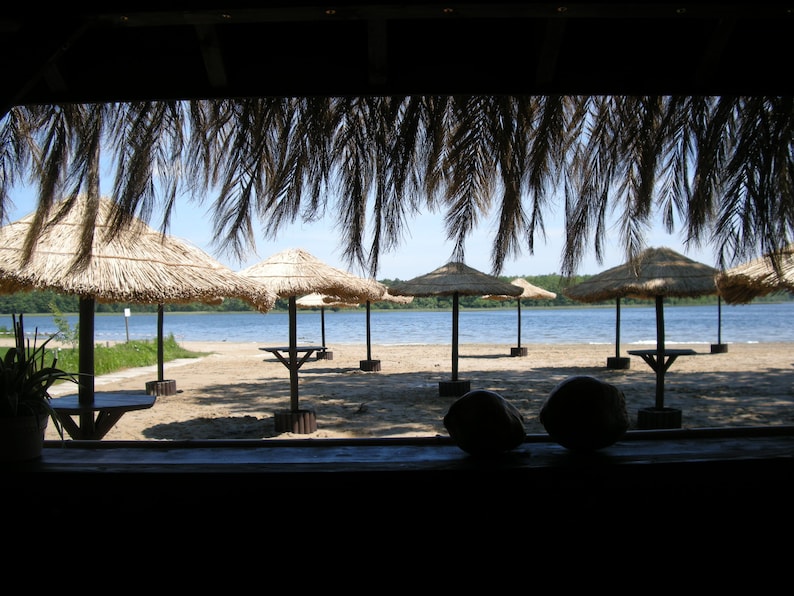 K&ouml;nnte beinhalten: Eine Strandlandschaft mit mehreren Strohschirmen, die Schatten spenden. Die Schirme sind mit Tischen aufgestellt und blicken auf einen ruhigen See unter einem klaren blauen Himmel. Die Szene wird von einem Strohdach eingerahmt.