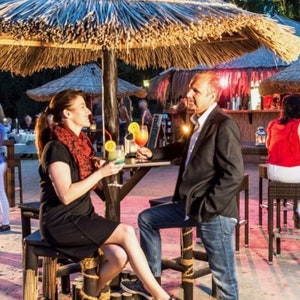 K&ouml;nnte beinhalten: Szene einer Outdoor-Bar mit Menschen, die Cocktails genie&szlig;en. Tische und Hocker stehen unter Strohschirmen. Die Bar hat eine tropische Atmosph&auml;re mit Sandboden. Cocktails sind mit Fr&uuml;chten garniert.