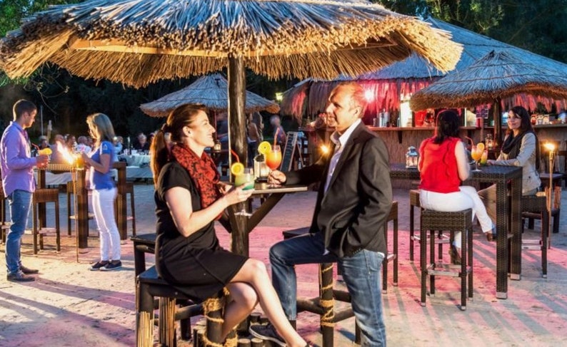 K&ouml;nnte beinhalten: Szene einer Outdoor-Bar mit Menschen, die Cocktails genie&szlig;en. Tische und Hocker stehen unter Strohschirmen. Die Bar hat eine tropische Atmosph&auml;re mit Sandboden. Cocktails sind mit Fr&uuml;chten garniert.