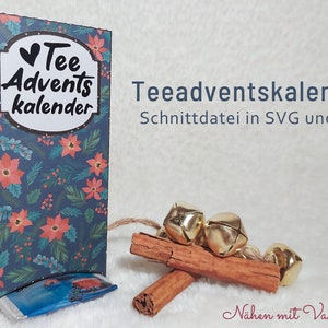 Op de afbeelding: Een tealblauwe doos met een rood en groen bloemenpatroon. De doos heeft een wit label met de tekst "Tee Adventkalender" en een hart. De doos staat op een wit oppervlak met kaneelstokjes, gouden belletjes en touw.