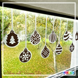 May include: A window with a view of a green lawn and trees decorated with brown and white Christmas baubles. The baubles are hanging from strings and have different designs, including snowflakes, stars, and a Christmas tree.