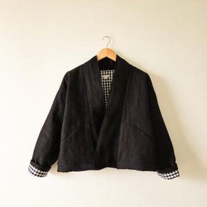 May include: A black jacket with a relaxed fit, hanging on a wooden hanger. The jacket features a contrasting gingham pattern on the inner lining and sleeve cuffs. The jacket has two front pockets and a collarless design.