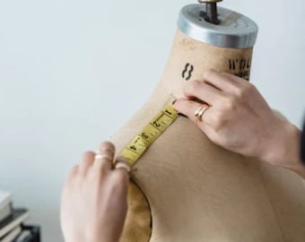 Puede incluir: Una persona mide el hombro de un maniquí de costura con una cinta métrica amarilla. El maniquí está hecho de tela beige y tiene un soporte de madera.