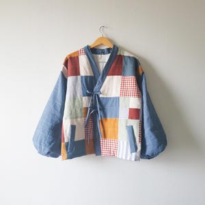 Op de afbeelding: Een patchworkjasje met een V-hals en striksluiting. De jas heeft een verscheidenheid aan stoffen vierkanten in kleuren als blauw, rood, oranje en crème. De mouwen zijn effen blauw en de jas hangt aan een houten hanger.