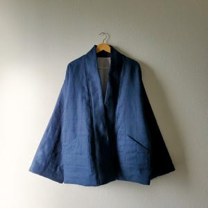 Puede incluir: Chaqueta azul marino estilo kimono con un patrón acolchado y dos bolsillos delanteros. La chaqueta cuelga de una percha de madera contra una pared de color neutro. La chaqueta tiene un cuello ancho y un corte holgado.