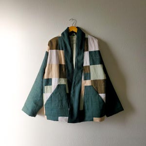 May include: A quilted jacket in shades of green, beige, and white hangs on a wooden hanger. The jacket features a patchwork design with square and rectangular patterns. The sleeves and front pockets are a solid teal green.
