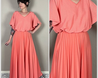 May include: A coral pink maxi dress with a pleated skirt and a short-sleeved top with a V-neckline. The top has a textured fabric and the sleeves have ties at the shoulders.