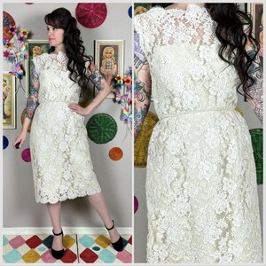 May include: A vintage ivory lace dress with a high neckline and cap sleeves. The dress features a fitted bodice and a knee-length skirt, cinched at the waist with a thin belt. The intricate floral lace pattern covers the entire dress.