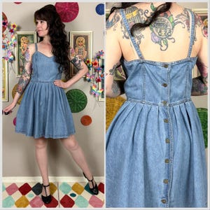 May include: A light blue denim dress featuring a sweetheart neckline, thin shoulder straps, and a pleated skirt. The back of the dress includes a vertical row of six metal buttons running from the top edge down to the waistline seam.