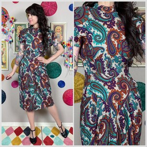 Puede incluir: Un vestido vintage con un vibrante estampado de cachemira en verde azulado, morado y naranja sobre un fondo crema. El vestido tiene mangas cortas y un cuello alto. El vestido llega a la rodilla y tiene un corpiño ajustado.