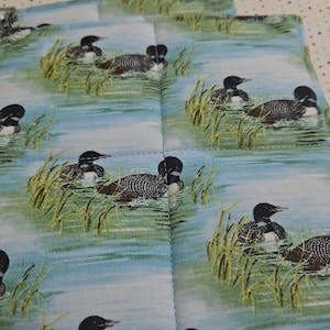 May include: Square quilted potholders with a repeating pattern of loons swimming in a lake. The loons have black heads and white-speckled bodies. The background is a light blue water with green reeds.